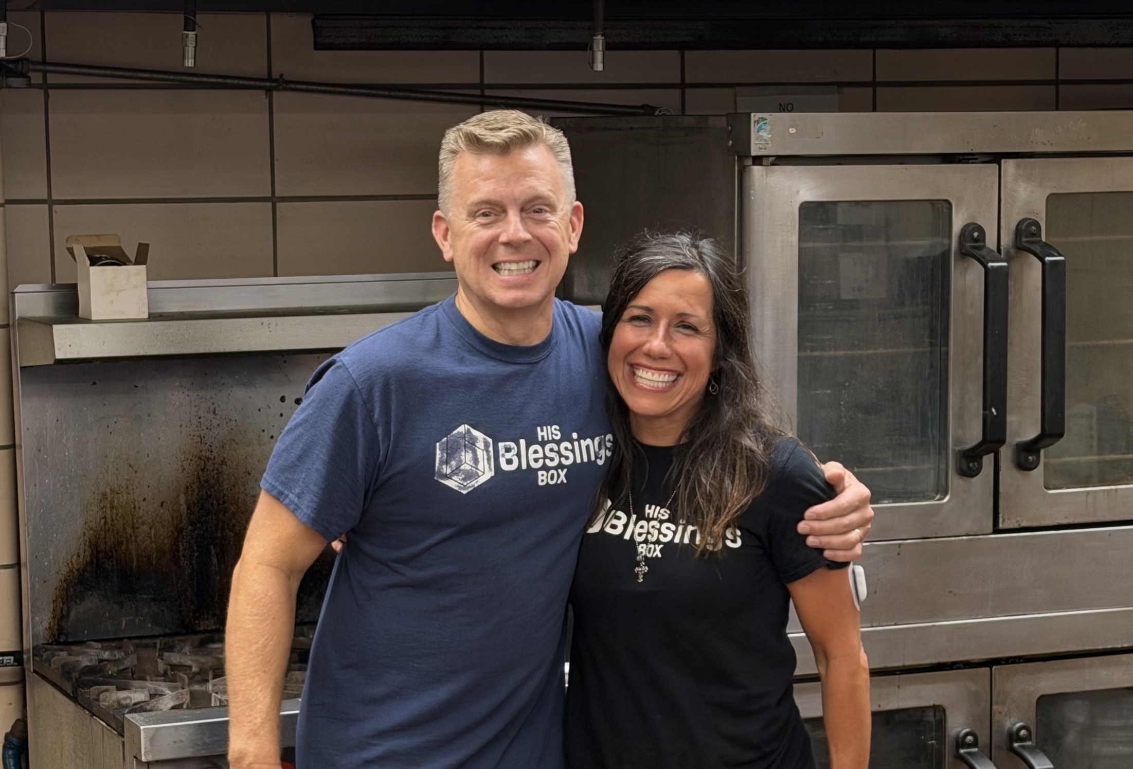 Two smiling volunteers in His Blessings Box shirts standing arm in arm in front of the kitchen ovens